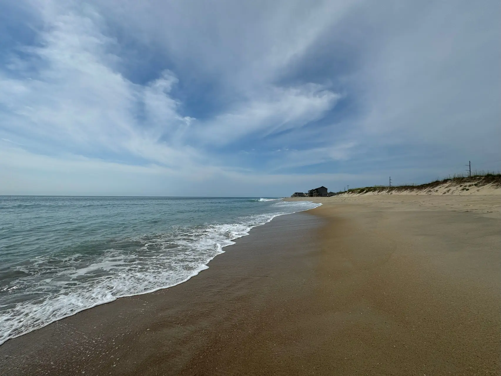 Wide sandy beach on the North Carolina Outer Banks with ocean surf, dunes, and distant beach houses under a partly cloudy sky
